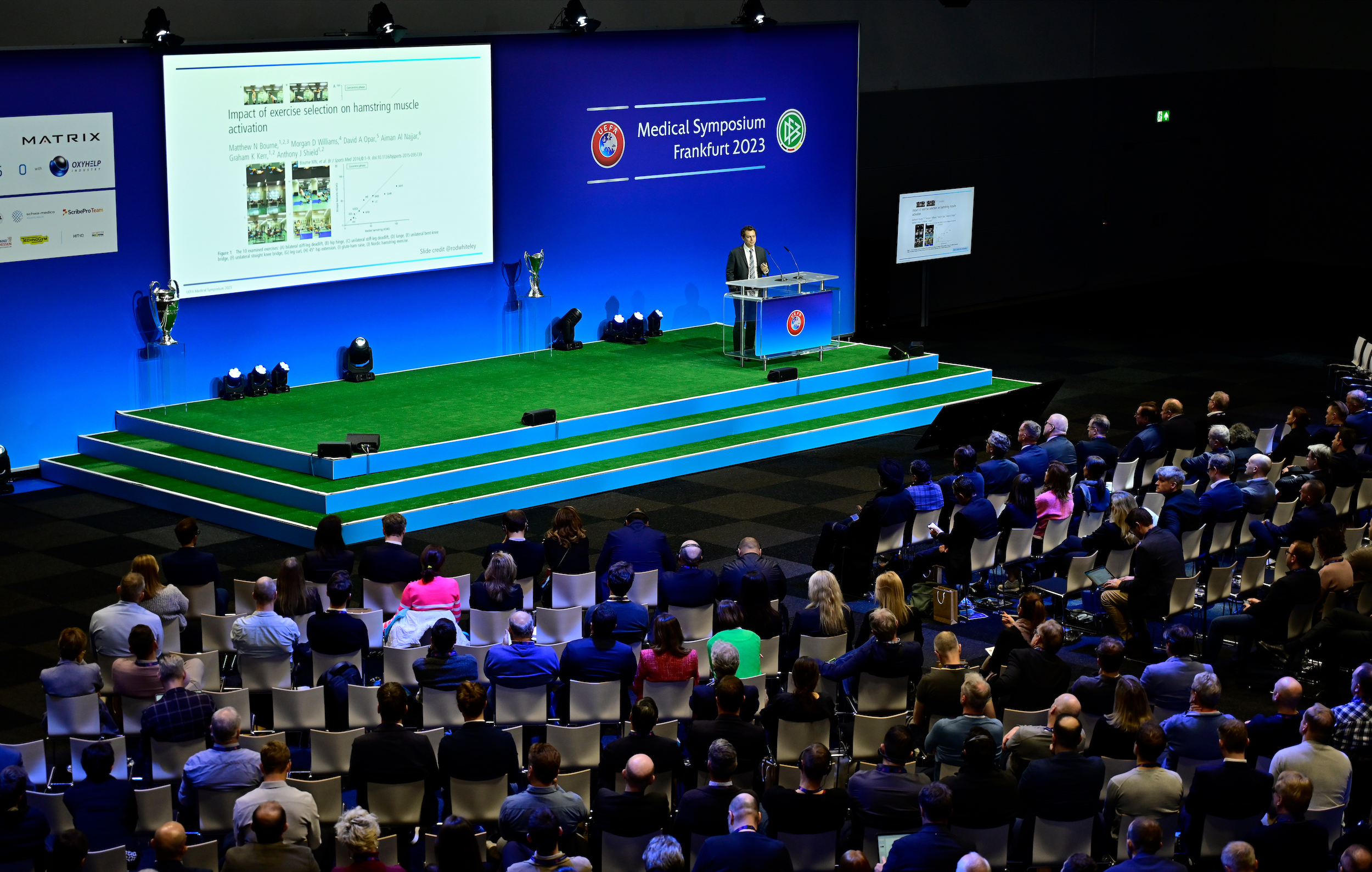The picture shows the stage of the UEFA Medical Symposium. A speaker is giving a lecture. 